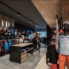Ein Bekleidungsgeschäft mit Holzdecke und gefliestem Boden. Verschiedene Kleidungsstücke hängen an den Kleiderstangen und einem Holzregal. Ein Mannequin ist in orangefarbener Hose und einer grauen Jacke mit einer weißen Mütze gekleidet.