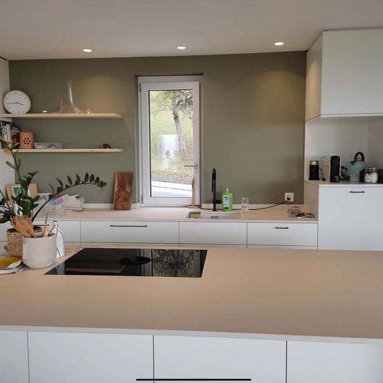 Eine moderne Küche mit weißen Schränken und einer beigen Arbeitsplatte. Die Küche verfügt über eine Glastür, eine wandmontierte Uhr und Regale mit Dekorationsartikeln.