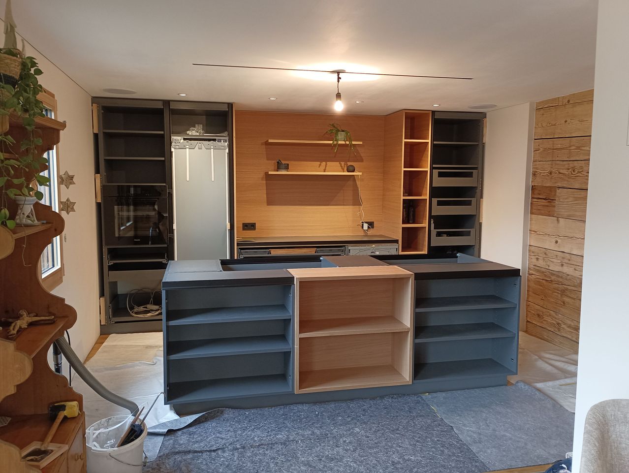 Eine unfertige Küche mit grauer Arbeitsplatte, Holzregalen und Schränken. Im Hintergrund befinden sich ein Kühlschrank, ein Herd und ein Waschbecken. Ein Eimer und ein Schlauch liegen auf dem Boden.