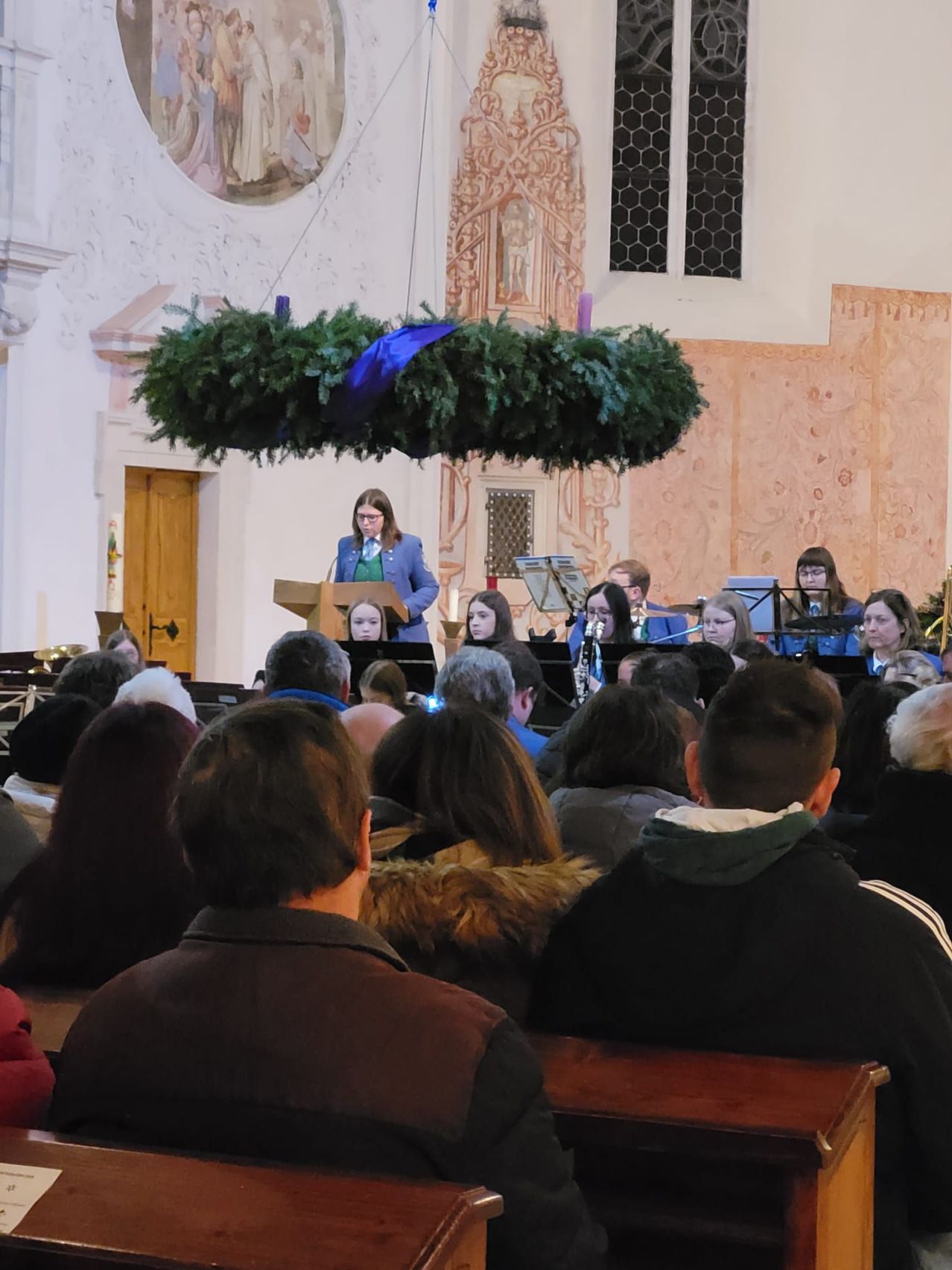 Eine Frau spricht an einer Kanzel in einer Kirche, während eine Band im Hintergrund spielt. Das Publikum sitzt vor ihnen.