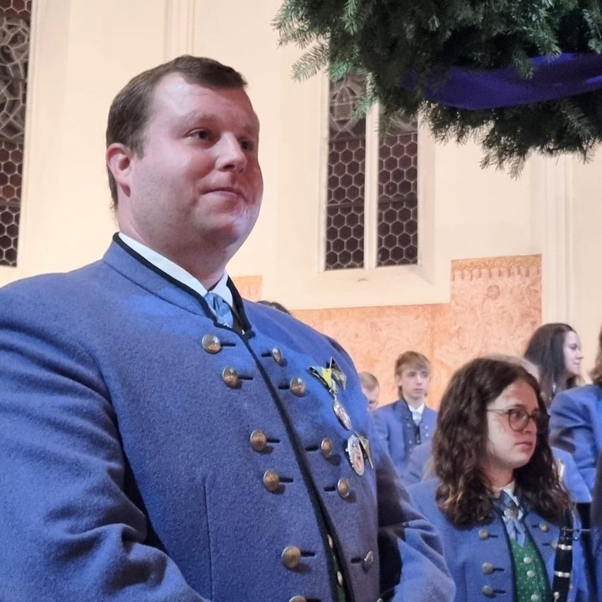 Ein Mann in einem blauen Uniform steht in einer Kirche, möglicherweise während einer Zeremonie. Neben ihm hält eine Frau in einer ähnlichen Uniform eine Klarinette. Hinter ihnen sind mehrere andere Personen in Uniformen anwesend, und ein Kranz hängt an der Wand.