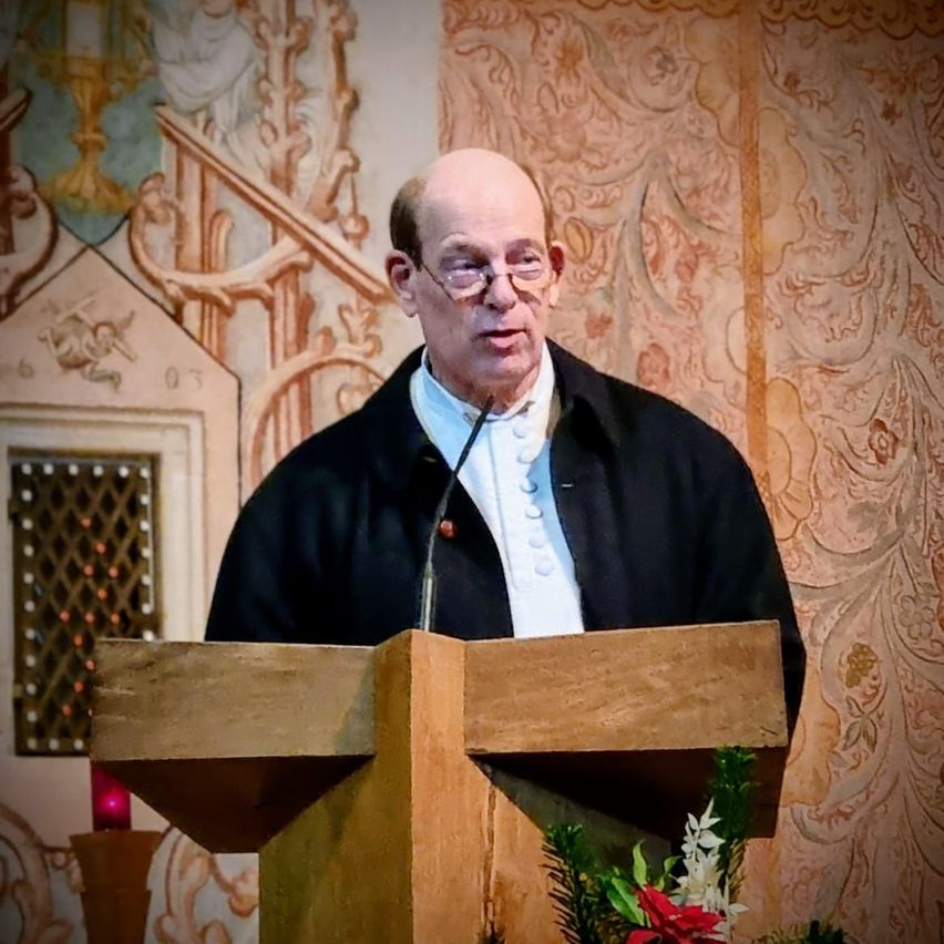 Ein älterer Mann mit Brille und schwarzem Gewand steht an einem hölzernen Rednerpult mit Mikrofon und hält eine Rede in einer Kirche.