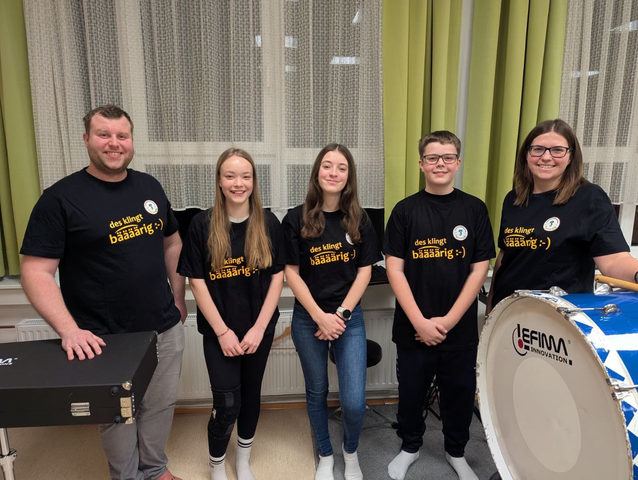 Five people are standing together in front of a drum and smiling. They all wear black T-shirts with yellow writing on them. The man on the left holds a black box.