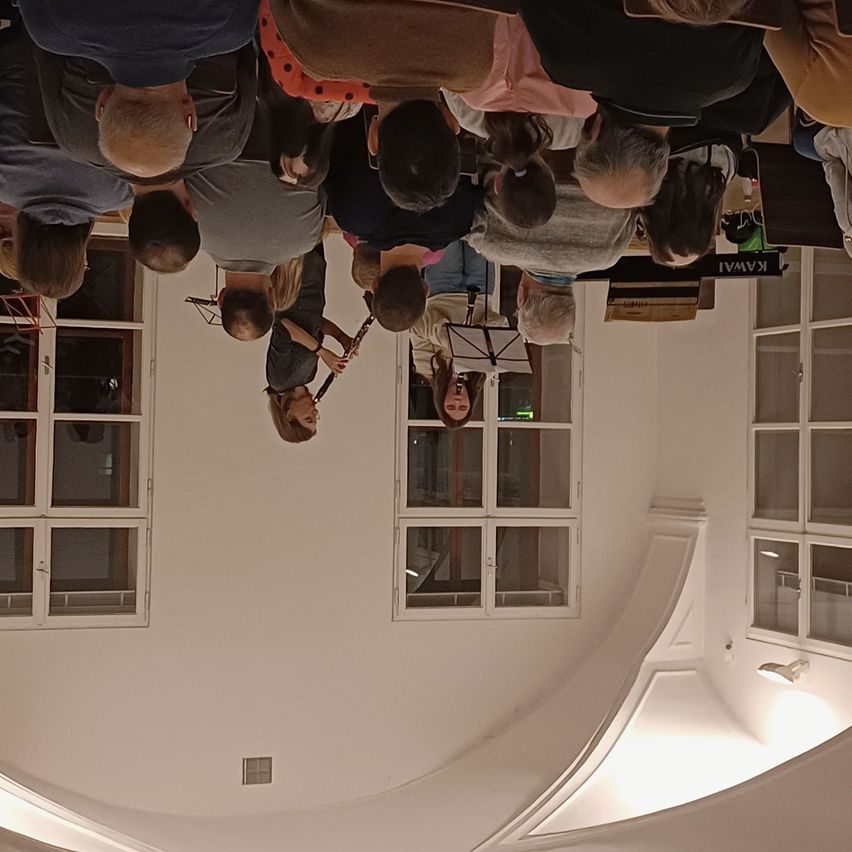 Eine Gruppe von Menschen versammelt sich in einem Raum und spielt Musikinstrumente. Eine Frau spielt Klarinette, während andere ein Saxophon spielen. Im Raum befinden sich Fenster und ein helles Licht.