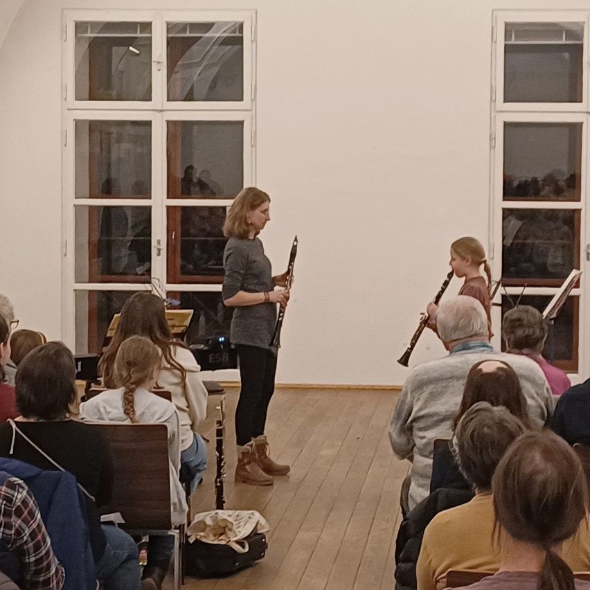 Eine Frau und ein Mädchen spielen Klarinette in einem Raum mit Holzboden, weißen Wänden und großen Fenstern. Mehrere Menschen sitzen vor ihnen und hören aufmerksam zu.