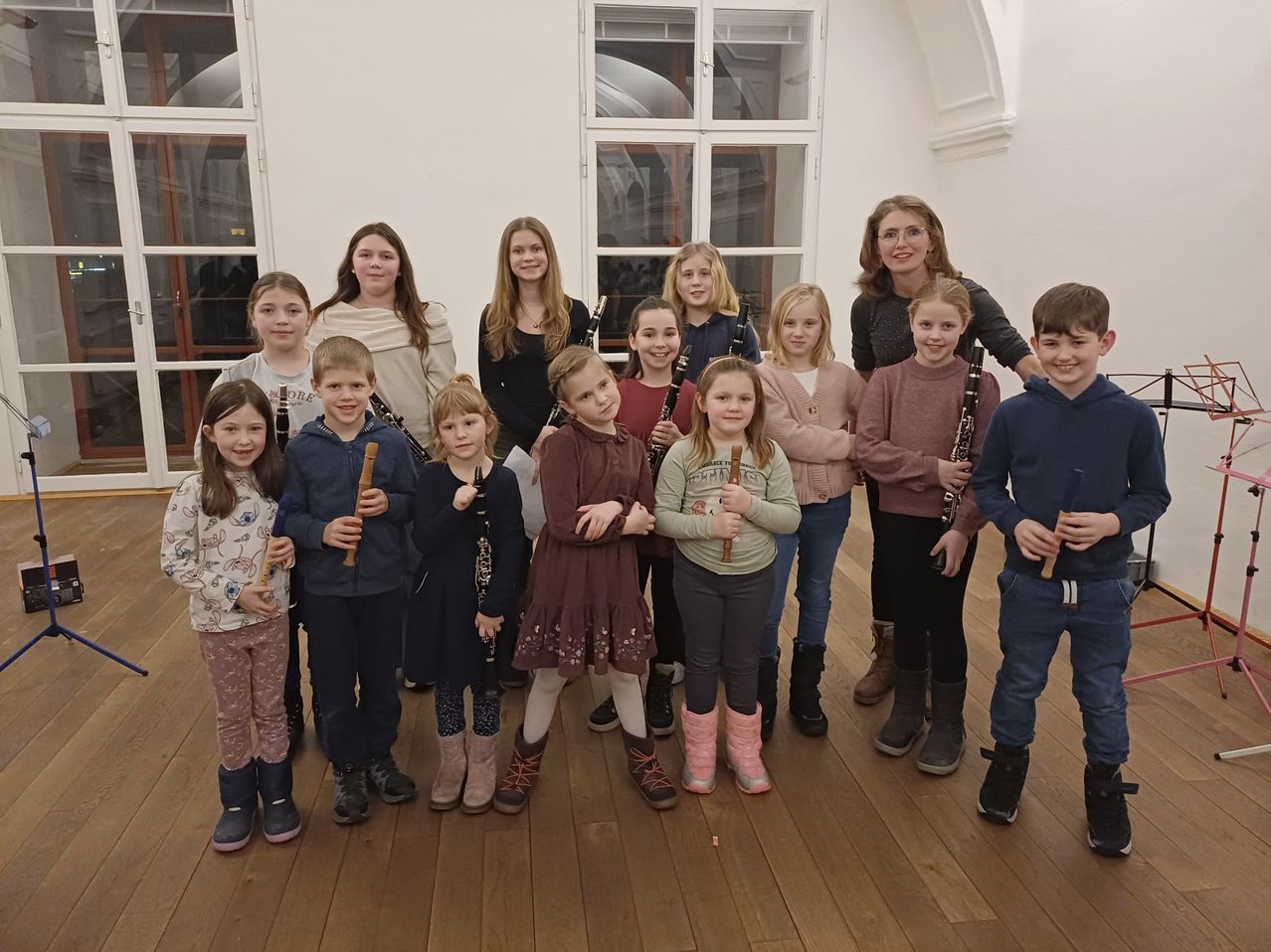 Eine Gruppe von Kindern mit Musikinstrumenten, möglicherweise Klarinetten, posiert für ein Foto in einem Raum mit Holzboden und Glasfenstern.