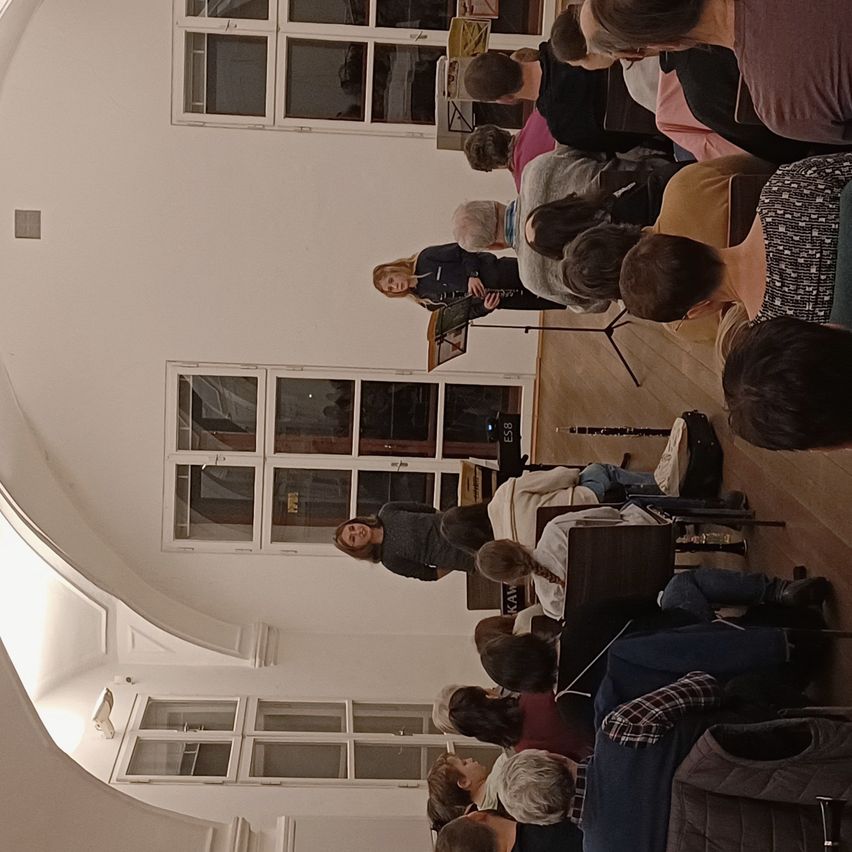 Ein Innenbereich mit gewölbter Decke, eine Frau steht in der Mitte und spielt die Flöte, mehrere Menschen sitzen vor ihr, und es gibt große Fenster.