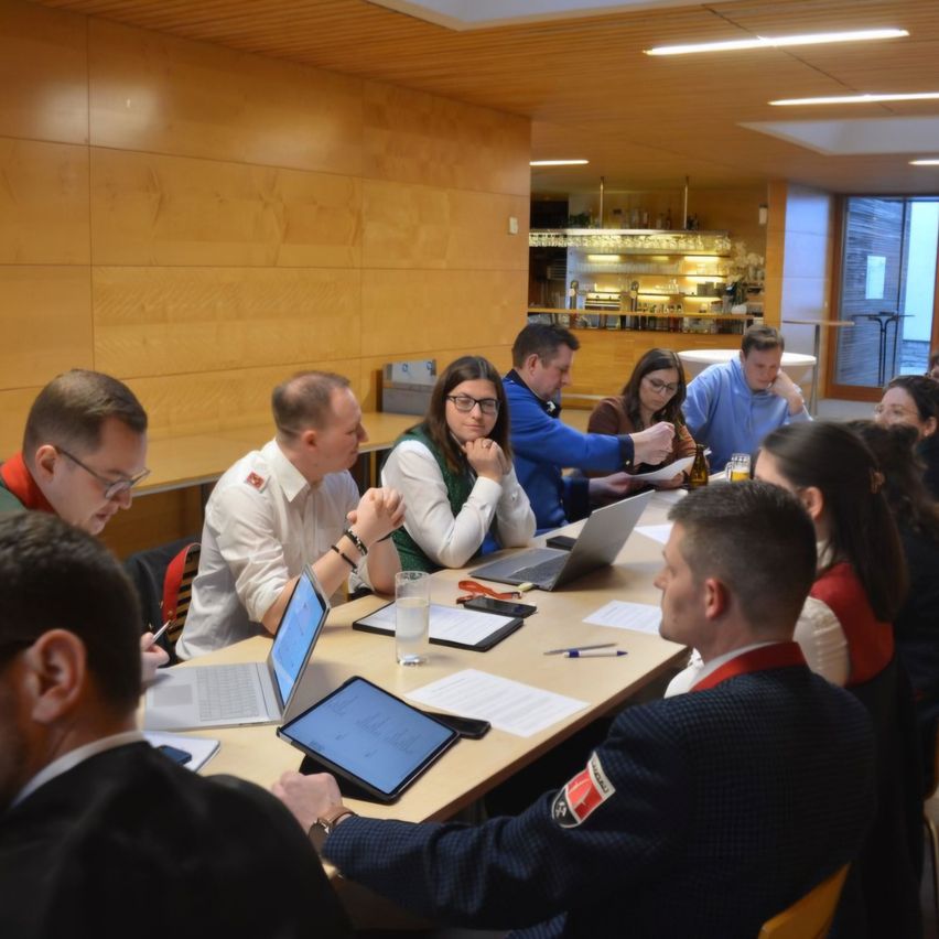 A group of people are sitting around a table with laptops and papers, likely in a meeting. The room has wooden walls and a ceiling with lights. There are also chairs and a glass window.