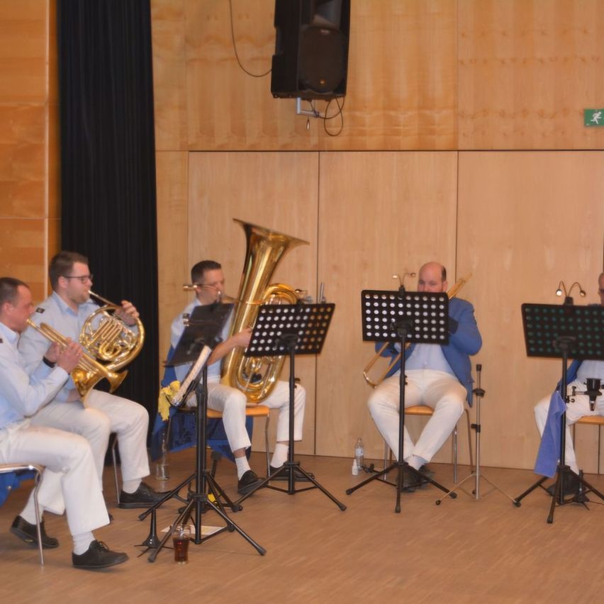 Five men dressed in matching white pants and blue shirts are playing brass instruments. They sit on chairs with music stands in front of them. A speaker is mounted on the wall behind them.