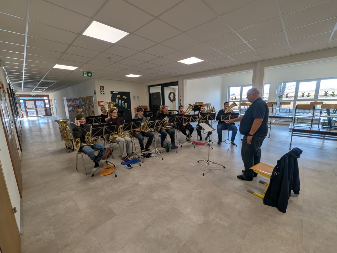 Eine Gruppe von Musikern übt in einer Halle mit gefliesten Böden und weißen Decken. Sie sitzen in Reihen vor einem Mann, der möglicherweise ihr Lehrer ist.