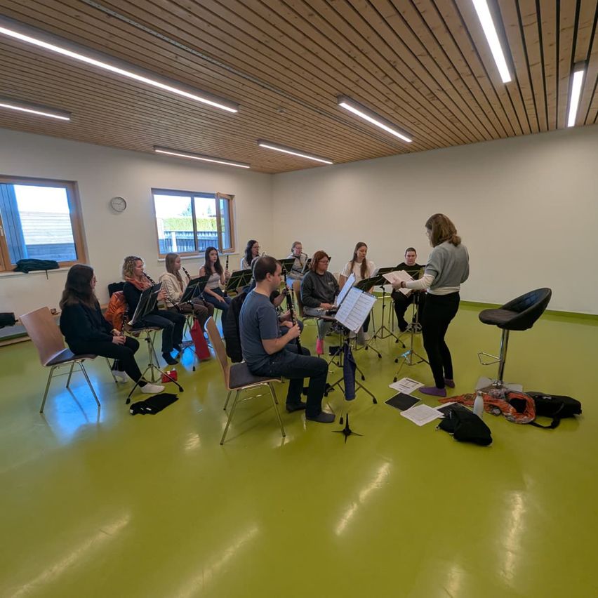 Eine Gruppe von Musikern ist in einem Raum mit grünem Boden versammelt. Sie sitzen und spielen verschiedene Instrumente. Eine Frau steht vor ihnen und hält Notenblätter. Der Raum hat weiße Wände, eine Holzdecke und große Fenster.