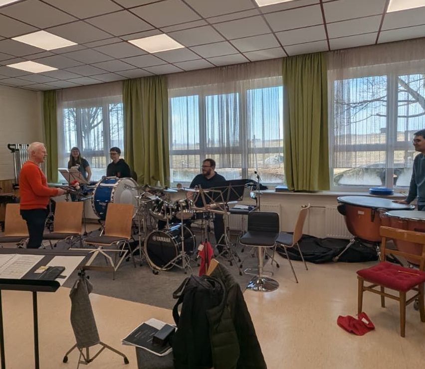 In einem Raum mit großen Fenstern versammeln sich mehrere Menschen um ein Schlagzeug, um zu üben. Der Raum hat weiße Wände, einen weißen Heizkörper und grüne Vorhänge. Stühle und Musikinstrumente sind verstreut.