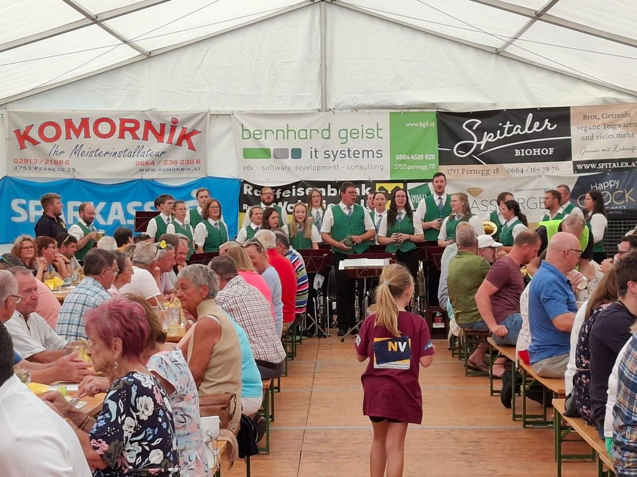 A choir performs on stage under a tent, with a diverse audience seated in front. Banners with various logos and text are displayed behind the choir.