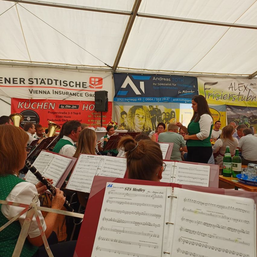 A brass band is performing under a tent, with musicians playing various instruments. A woman stands in front, possibly conducting. Banners with logos and text are visible in the background.