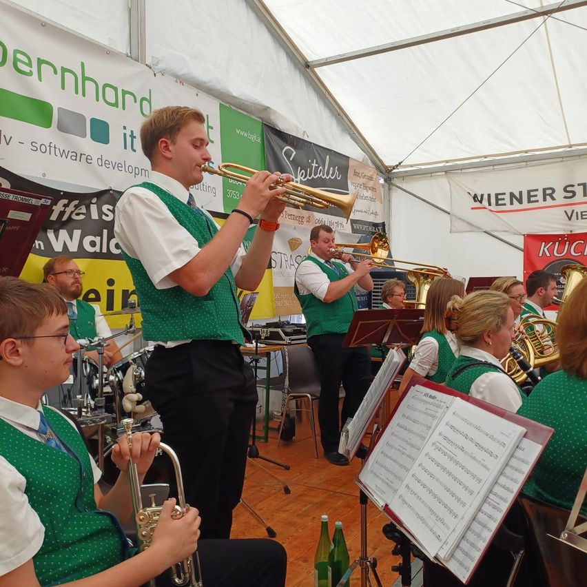 A group of musicians in green vests play brass instruments in a tent, with a man playing the trumpet in the foreground and others playing trombones and tubas.