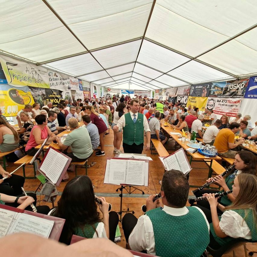 A large group of people are seated at long tables in a tent, with a band playing in the center. The band members are dressed in green vests and playing clarinets. The audience is enjoying the performance.