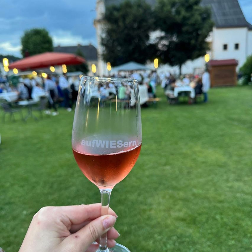 Eine Hand hält ein Weinglas mit rosa Wein vor einem verschwommenen Outdoor-Event mit Menschen, die an Tischen sitzen, und einem großen Gebäude im Hintergrund.