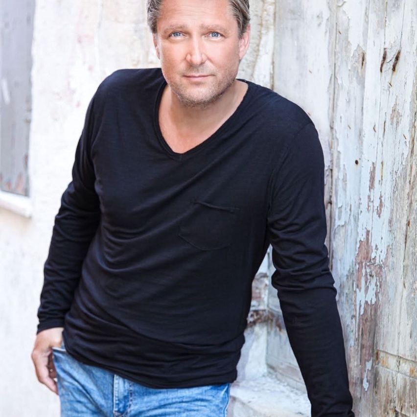 Ein Mann mit kurzem Haar und Bart steht vor einer weißen Wand mit abblätternder Farbe, trägt ein schwarzes Langarmshirt und blaue Jeans.