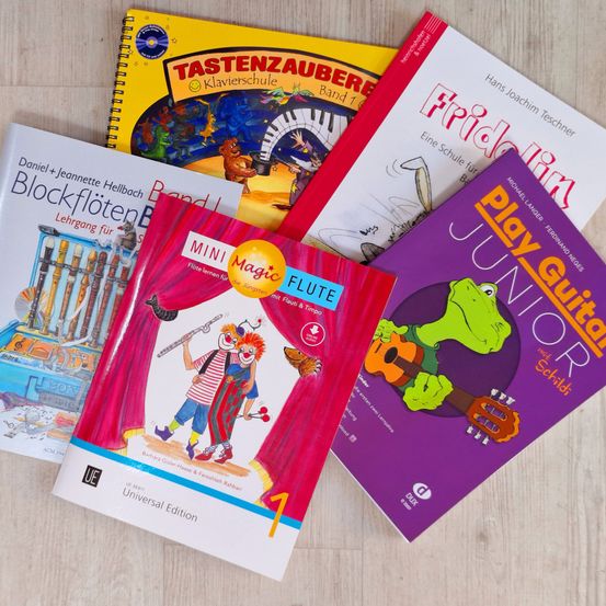 Fünf Bücher sind auf einer Holzoberfläche ausgestellt. Die Bücher sind betitelt Tastenzauber, Blockflötenbuch, MINI Magic Flute, Friedolin und Play Guitar Junior. Jedes Buch hat einen anderen farbigen Einband und einzigartige Illustrationen.