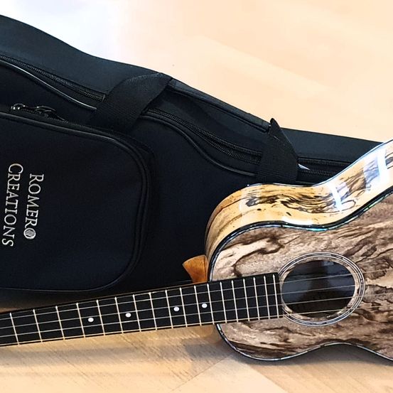 Ein braunes und schwarzes Ukulele liegt neben seiner schwarzen Tasche auf einem Holzboden. Die Ukulele hat ein einzigartiges Holzmuster auf ihrem Korpus. Die Tasche hat den Text 'ROMERO CREATIONS' darauf.