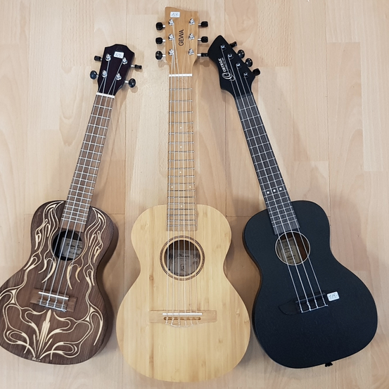 Drei Ukulelen sind auf einem Holzboden ausgestellt. Von links nach rechts gibt es eine braune Ukulele mit Flammendesign, eine hellbraune Ukulele und eine schwarze Ukulele.