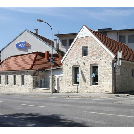 Ein Straßenblick mit einem kleinen Haus und einem blauen Schild mit der Aufschrift AM, einem Motorrad, das davor geparkt ist, einer Straßenlaterne und einem Gebäude im Hintergrund unter einem klaren blauen Himmel.