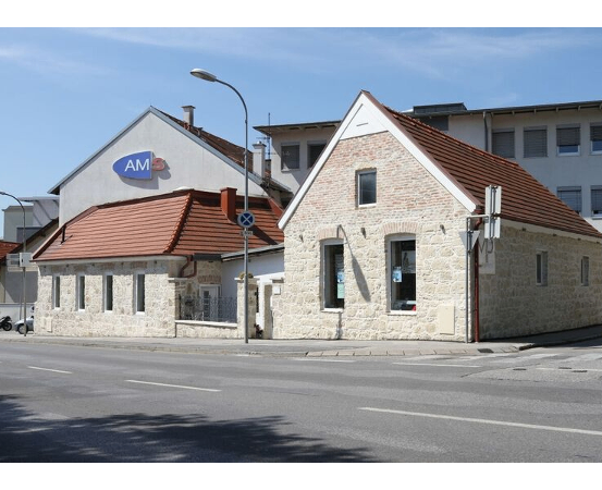 Ein Straßenblick mit einem kleinen Haus und einem blauen Schild mit der Aufschrift AM, einem Motorrad, das davor geparkt ist, einer Straßenlaterne und einem Gebäude im Hintergrund unter einem klaren blauen Himmel.