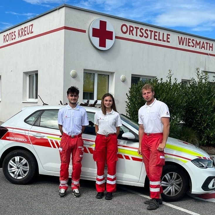 Three individuals in red and white uniforms stand in front of a white and red vehicle, likely an ambulance. They are in front of a building with a red cross sign.