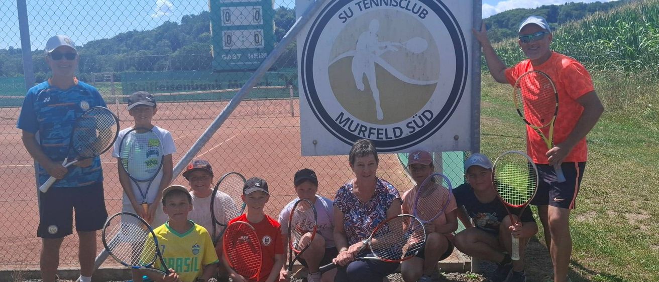 Eine Gruppe von Kindern und Erwachsenen posiert für ein Foto vor einem Tennisplatz mit einem Schild, das 'SU Tennisclub Murfeld Süd' steht.