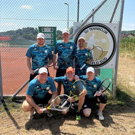 Fünf Männer posieren für ein Foto vor einem Tennisplatz. Sie tragen alle blaue Shirts und halten Tennisschläger. Dahinter steht ein Zaun mit einer Anzeigetafel und einem Schild mit einem Logo.