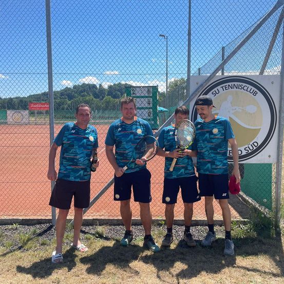 Vier Männer in passenden blauen Shirts und Shorts stehen gemeinsam auf einem Tennisplatz und halten Tennisschläger. Hinter ihnen befindet sich ein Tennisplatz mit einem Zaun und einem Schild, das 'SU Tennisclub' steht. Bäume und Berge sind im Hintergrund zu sehen.