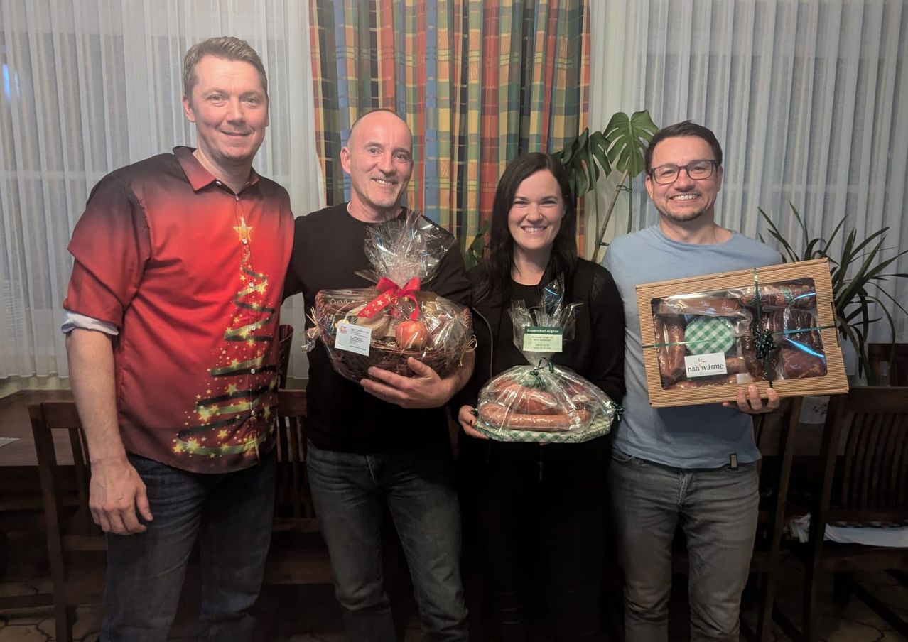 Vier Personen stehen in einem Raum. Sie halten Geschenkkörbe. Ein Mann in einem Weihnachtshemd steht links. Dahinter ist ein Vorhang.