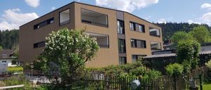 Ein modernes braunes Gebäude mit mehreren Stockwerken und Fenstern. Es hat einen kleinen Garten mit einem grünen Tisch und Stühlen. Das Gebäude ist von einem Holzzaun und Büschen umgeben.