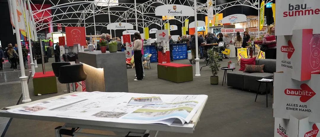 Eine Indoor-Ausstellung mit mehreren Ständen, einer grünen Bank, Topfpflanzen und Menschen, die herumlaufen. Eine Person steht vor einem Schreibtisch mit offenem Buch.