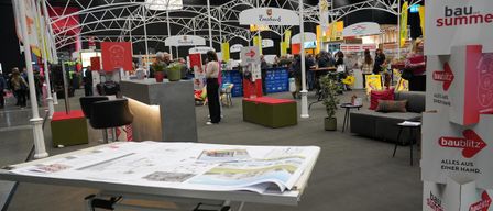 Eine Indoor-Ausstellung mit mehreren Ständen, einer grünen Bank, Topfpflanzen und Menschen, die herumlaufen. Eine Person steht vor einem Schreibtisch mit offenem Buch.