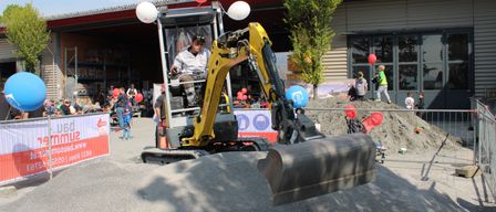 Ein Mann bedient einen gelben Bagger in einem öffentlichen Bereich. Es sind Menschen und rote Luftballons in der Nähe. Ein Schild mit Text ist im Vordergrund sichtbar.