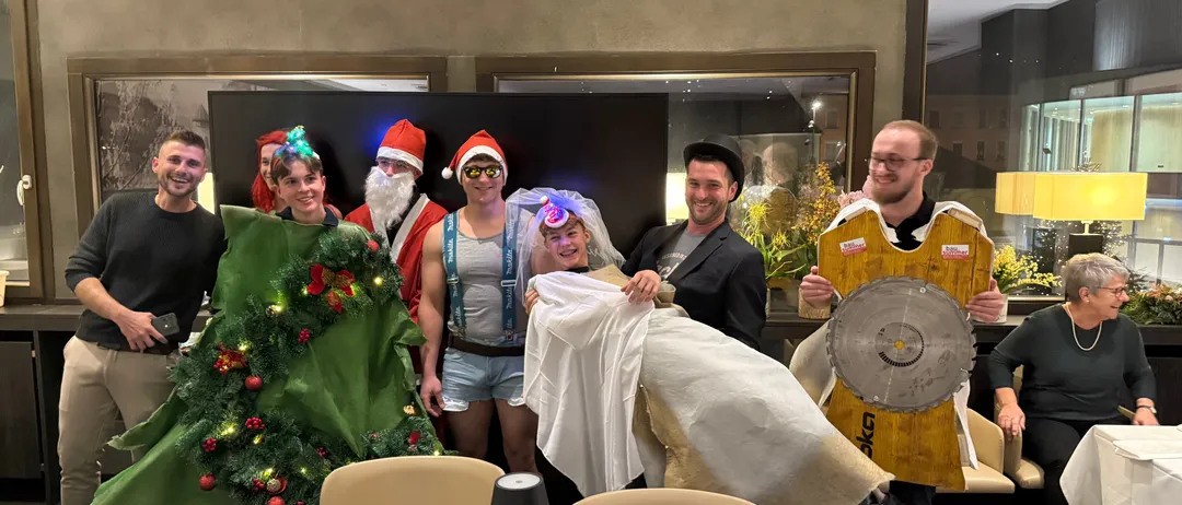 Eine Gruppe von Menschen in festlichen Kostümen posiert für ein Foto in einem Raum mit einem Weihnachtsbaum, Santa Claus und einer Säge.