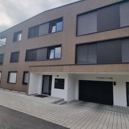 Ein modernes Gebäude mit brauner Holzfassade und Glasfenstern, ausgestattet mit Jalousien. Es hat einen gefliesten Eingang und eine schwarze Garagentür mit einem roten Teppich.