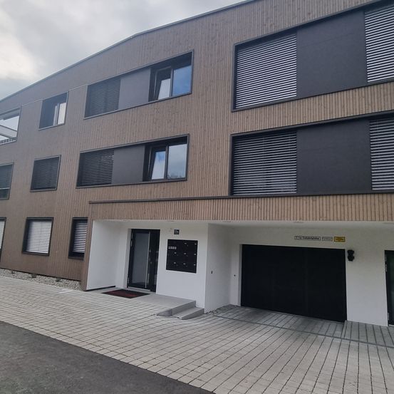 Ein modernes Gebäude mit brauner Holzfassade und Glasfenstern, ausgestattet mit Jalousien. Es hat einen gefliesten Eingang und eine schwarze Garagentür mit einem roten Teppich.