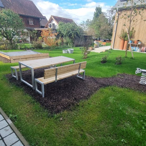 Ein Außenbereich mit einem Holztisch und einer Bank. Es ist von Gras umgeben mit einem Kiesweg. Es gibt Topfpflanzen und eine Spiralfeder auf dem Rasen.