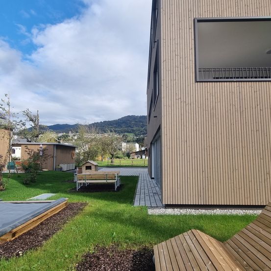 Ein modernes Gebäude mit Holzfassaden steht neben einem Rasen mit einer Bank, einem Hundehäuschen und einem Weg. In der Ferne befinden sich Berge unter einem bewölkten Himmel.