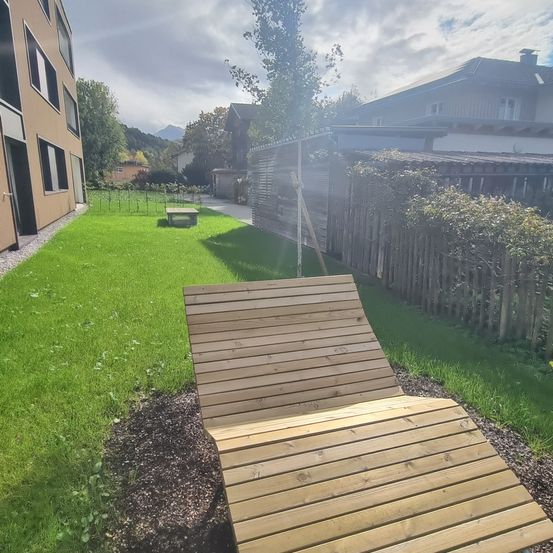 Eine Holzliege befindet sich in einem Garten mit Rasen, umgeben von einem Holzzaun und Gebäuden.