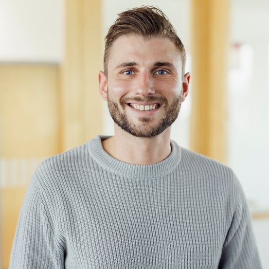 Ein Mann mit Bart und blauen Augen lächelt in die Kamera. Er trägt einen grauen Pullover und scheint sich in einem Büro zu befinden.