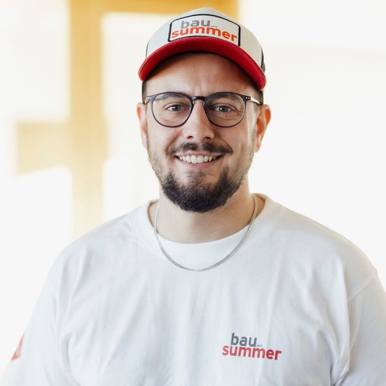 Ein lächelnder Mann in einem weißen T-Shirt mit 'bau mit summer' in Rot, trägt einen roten und weißen Hut. Er hat einen Bart und eine Brille.