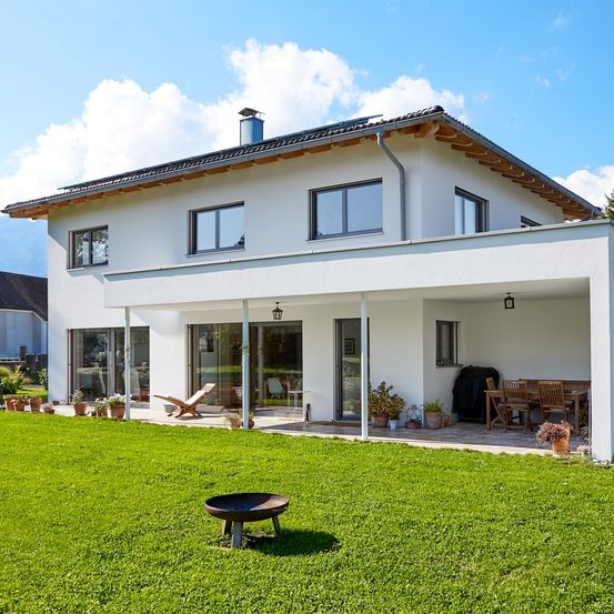 Ein modernes weißes Haus mit großen Fenstern und Glastüren, umgeben von einem gepflegten Garten mit einem Holztisch und Stühlen.
