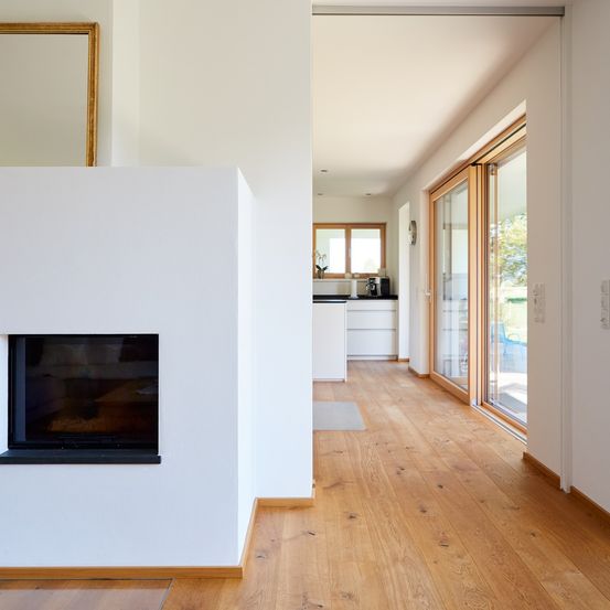 Ein leerer Flur mit Holzboden und weißen Wänden. Links befindet sich ein Kamin. Rechts führen Schiebetüren aus Glas zu einer Terrasse.