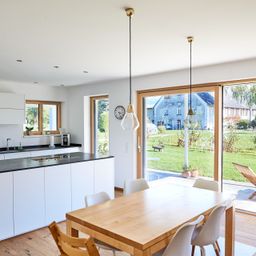 Ein moderner Essbereich mit einem Holztisch und weißen Stühlen. Eine weiße Küche mit schwarzem Arbeitsplatten und einem Waschbecken ist im Hintergrund. Der Raum hat eine weiße Decke mit Pendelleuchten. Glastüren führen in einen Garten mit Rasen und einem Haus.