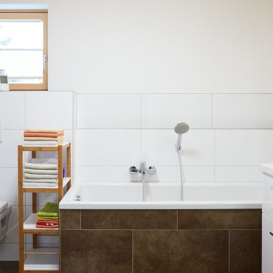 Ein Badezimmer mit weißen Wänden, einer Badewanne und Handtüchern auf einem Holzregal. Die Badewanne hat einen Wasserhahn und eine Duschbrause. Ein Fenster befindet sich links.