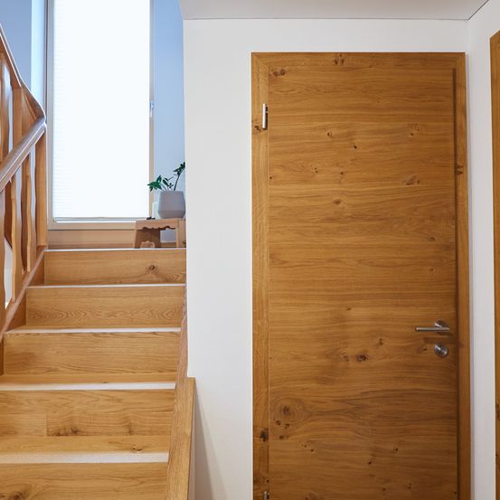 Eine Holztür neben einer Holztreppe, die zu einem Fenster mit Jalousien führt. Ein kleiner Topf mit einer Pflanze steht auf einem Holzhocker.
