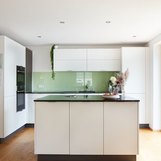 Eine moderne Küche mit weißen Schränken, einer grünen Glasrückwand und einer schwarzen Arbeitsplatte. Die Küche hat einen Waschbecken, einen Wasserhahn und eine Obstschale. Es gibt eine Steckdose und einen schwarzen Backofen an der Wand.